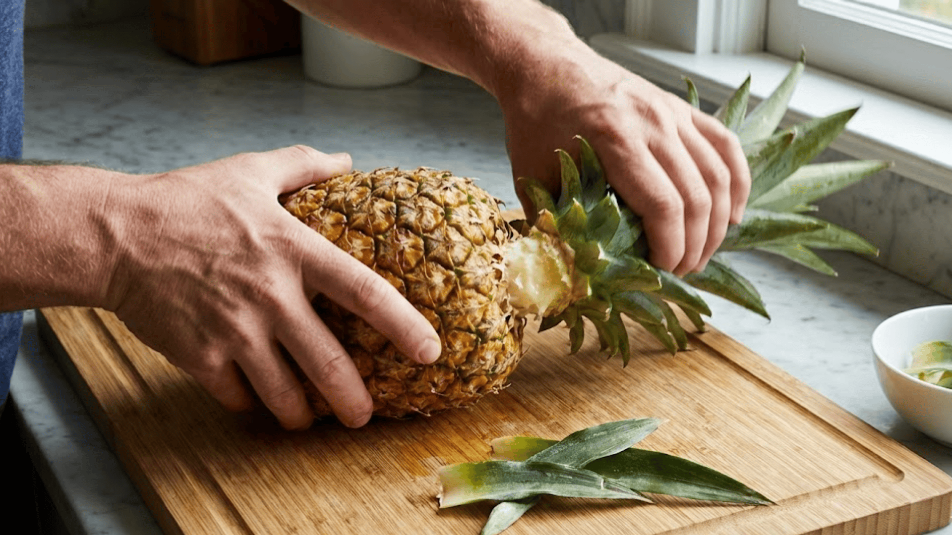 hands twisting pineapple crown off fruit on cutting board in bright kitchen