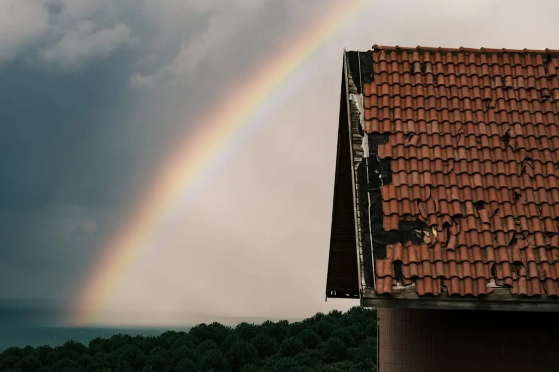 Rainbow arching over cloudy sky with partial view of red-tiled roof and trees