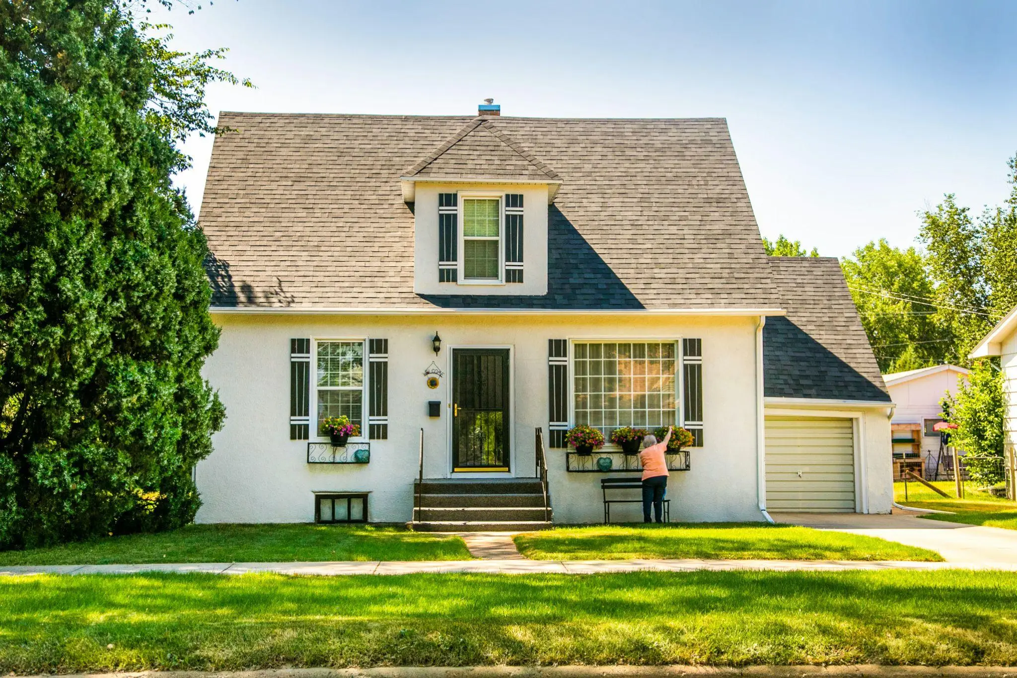 White house with gray roof and person tending flowers on the front porch in sunny weather