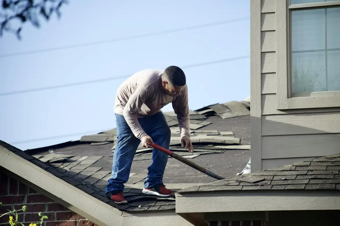 Worker repairing roof shingles on residential house under blue sky