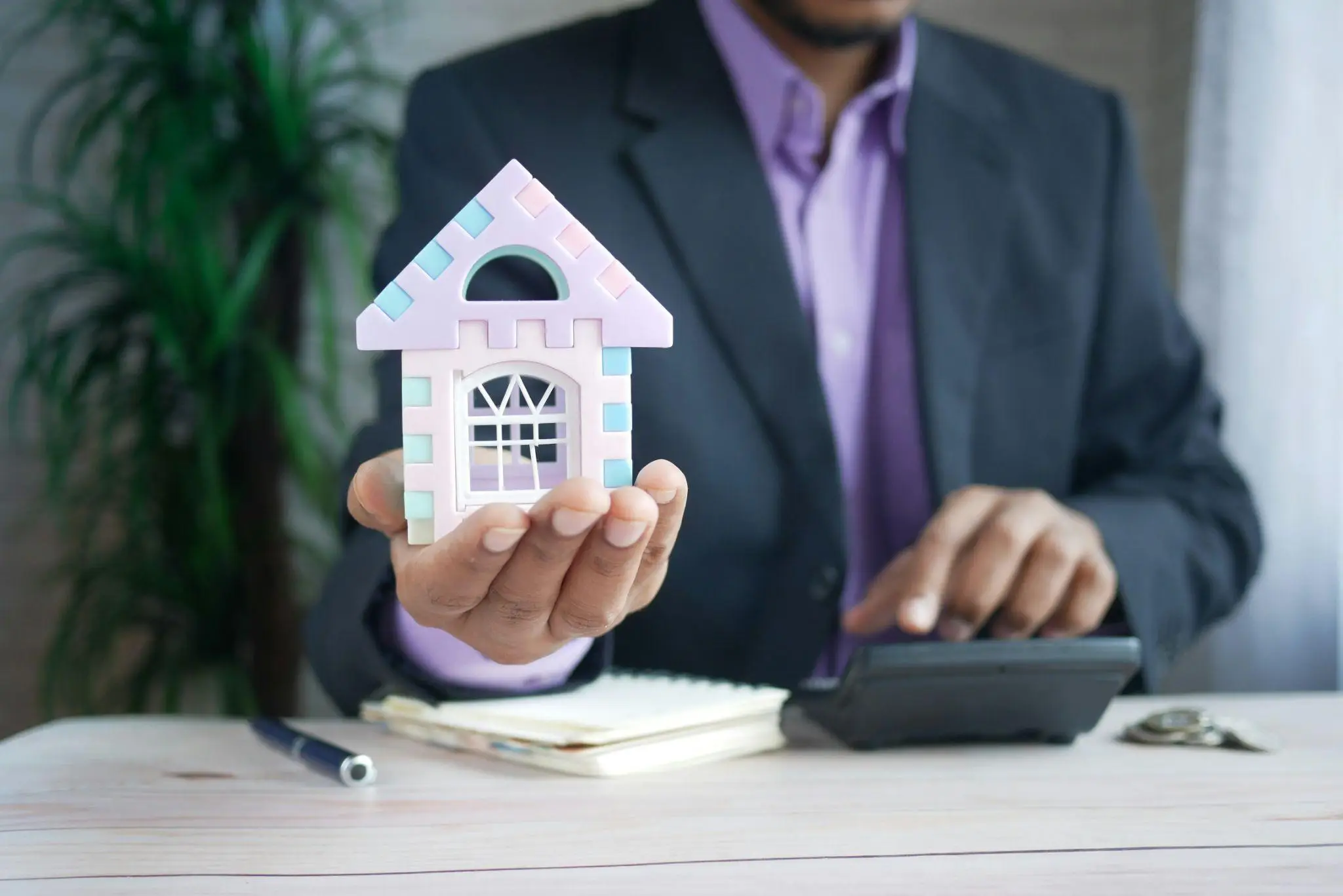Man in business suit holding toy house model while using calculator at desk