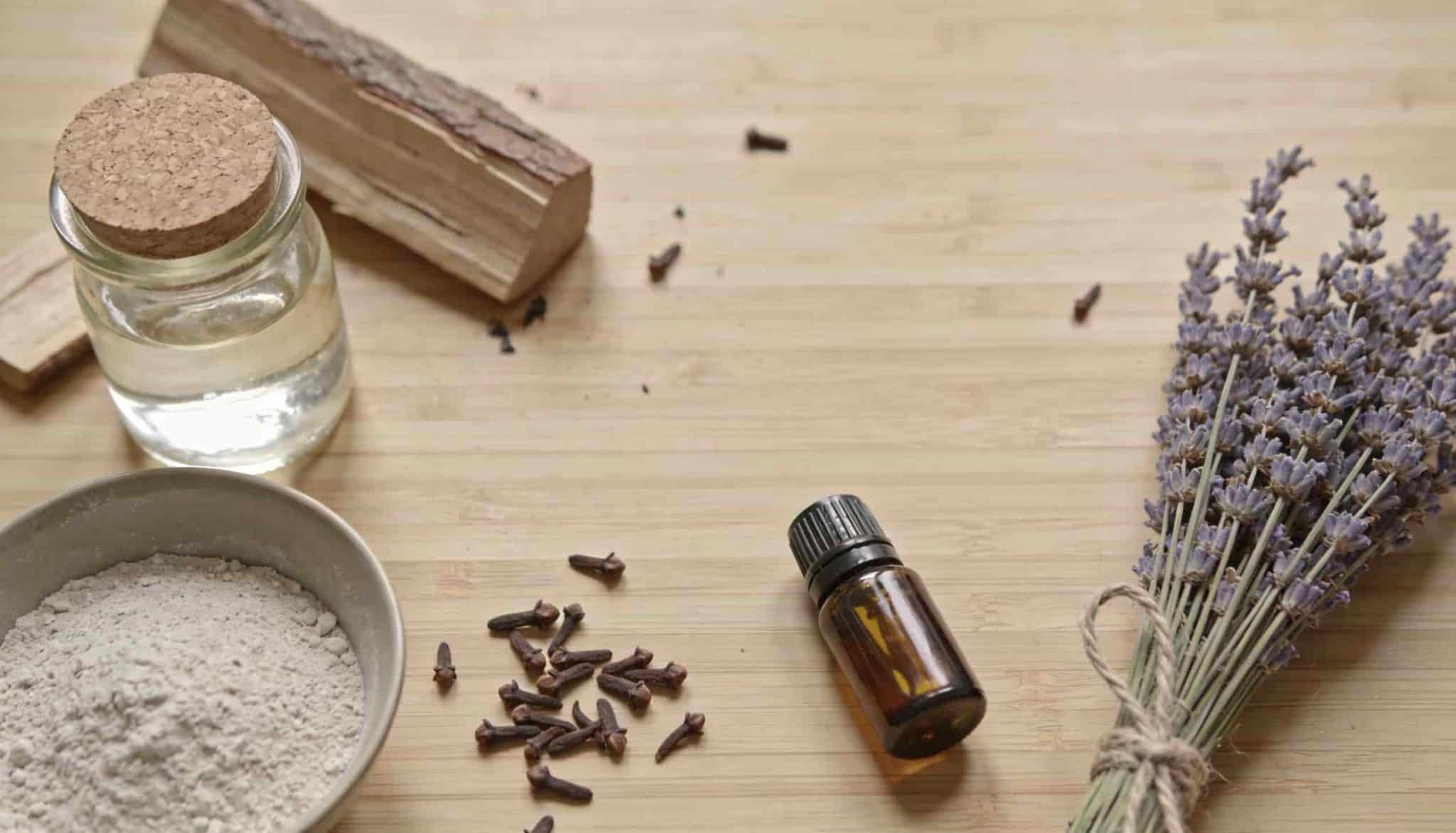 A wooden table holds natural pest control supplies, including a small glass jar, a bundle of dried lavender, and a small brown dropper bottle of essential oil.