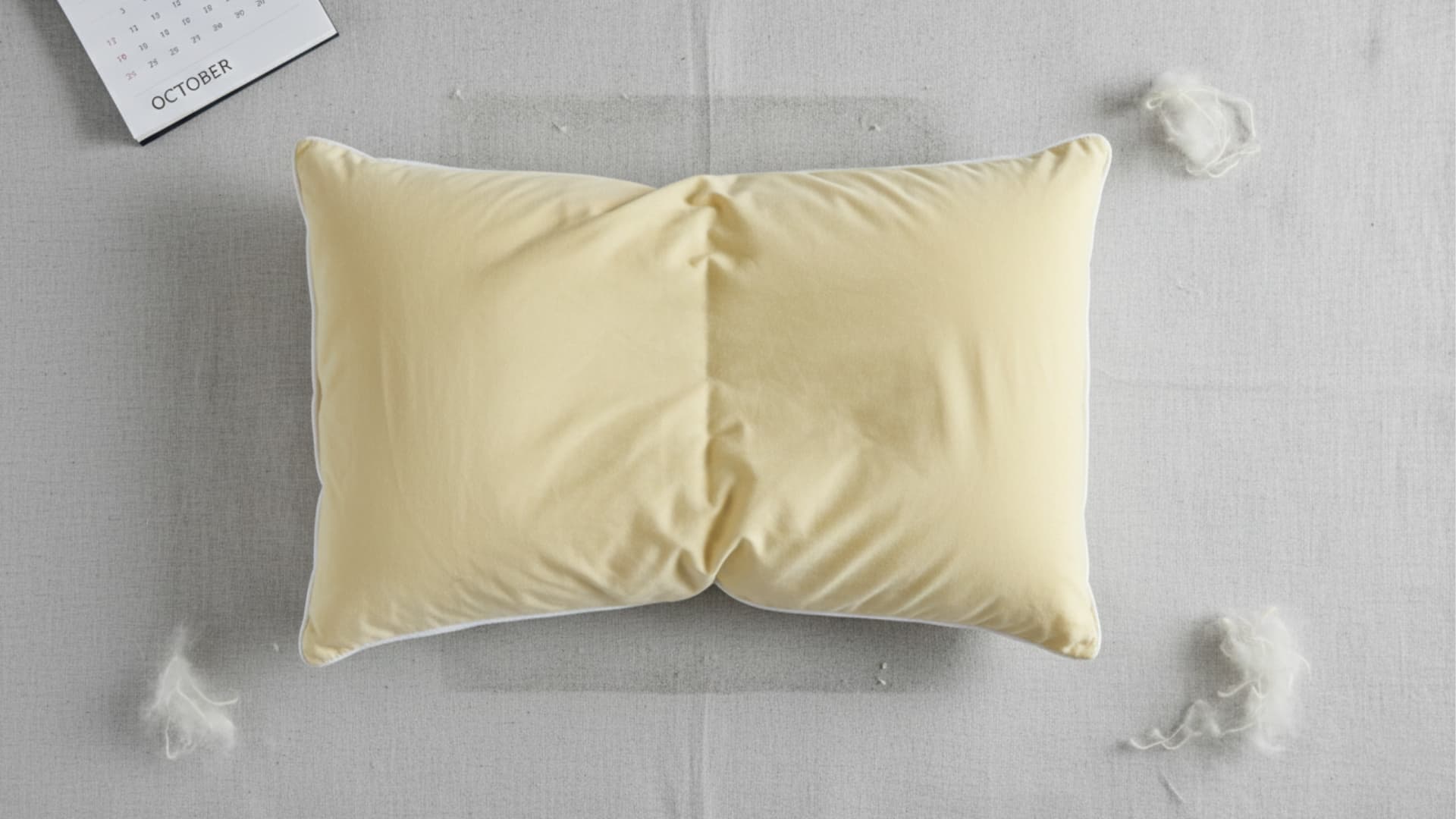 A worn, lumpy pillow with uneven filling lies on a surface, with loose stuffing around it, suggesting it needs replacement.