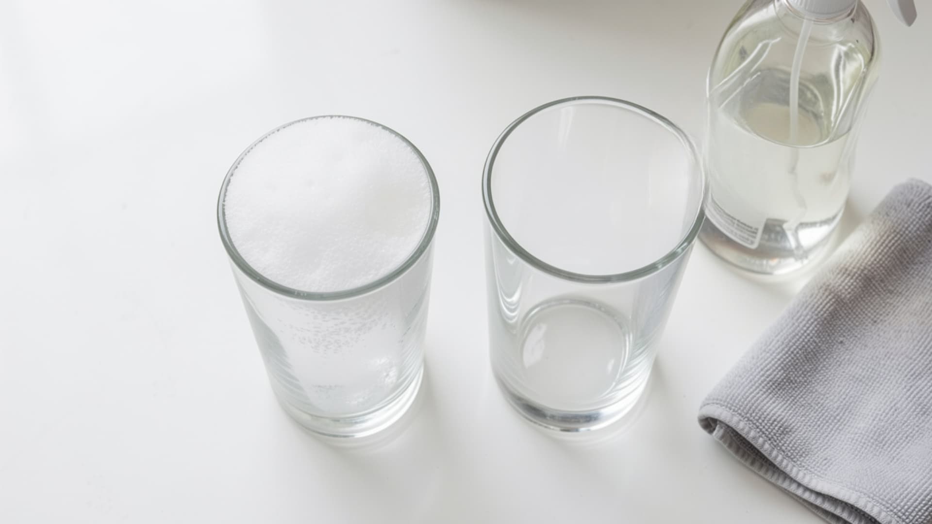 Glass with white hard water residue next to a clean empty glass, with a spray bottle and cloth nearby for cleaning glassware