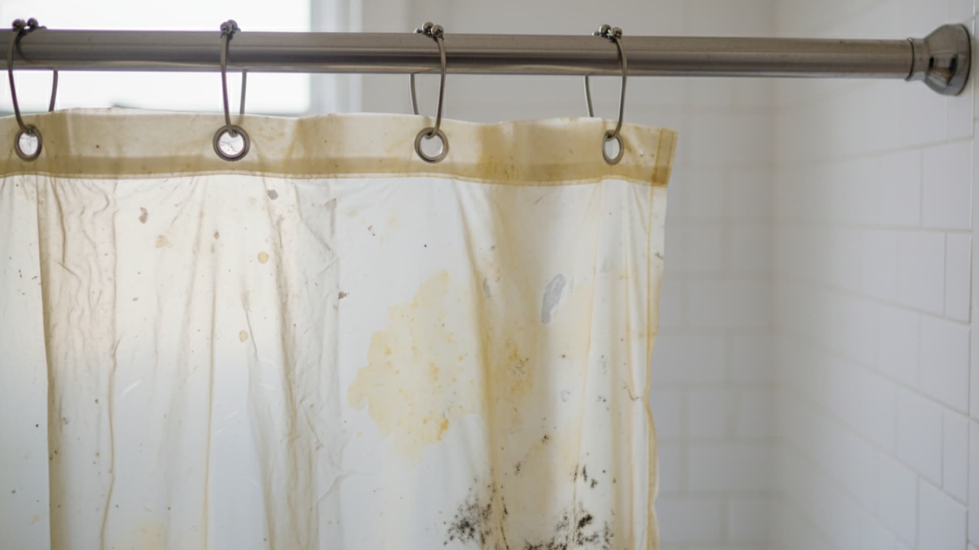 Old shower curtain hanging on a rod with visible yellow stains, mold spots, and discoloration indicating it needs replacement