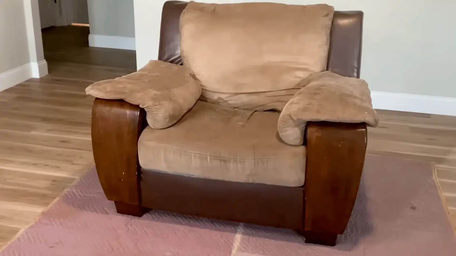 a beige fabric armchair with wooden accents and three cushions placed on a pink mat in a bright room