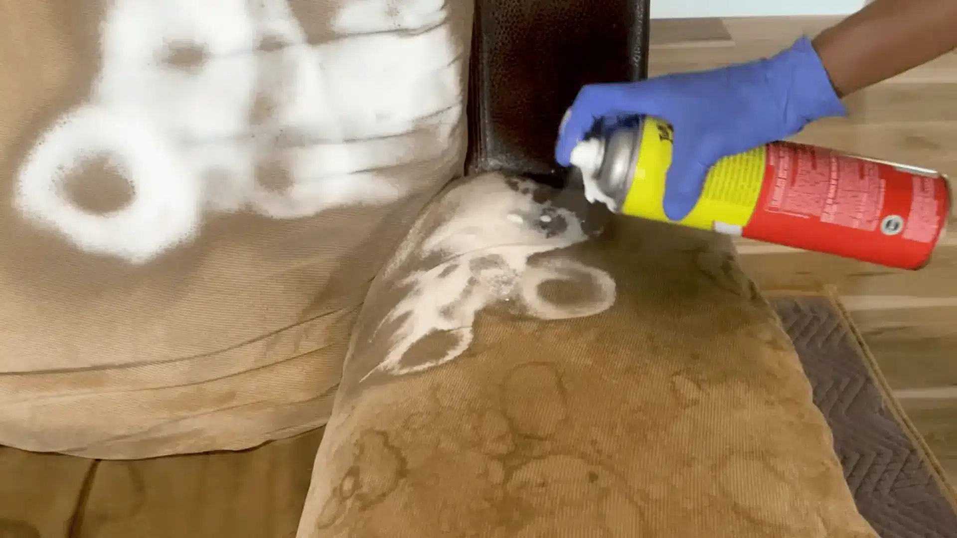 a gloved hand spraying foam cleaner onto a stained beige fabric couch cushion