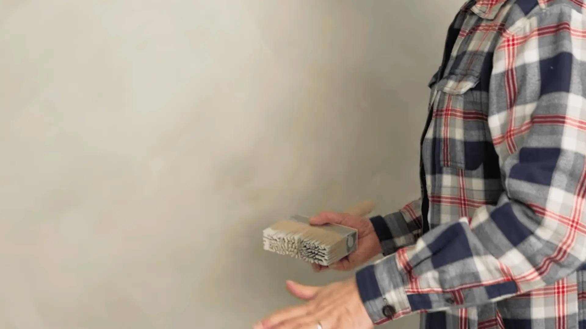 a person holding a stiff bristle brush in front of a completed limewash wall with a soft matte finish