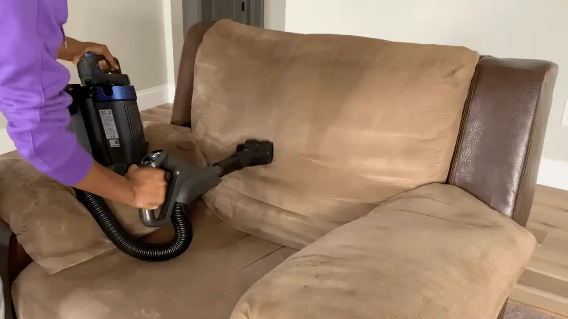 a person in a purple hoodie vacuuming a beige fabric couch with an upholstery attachment