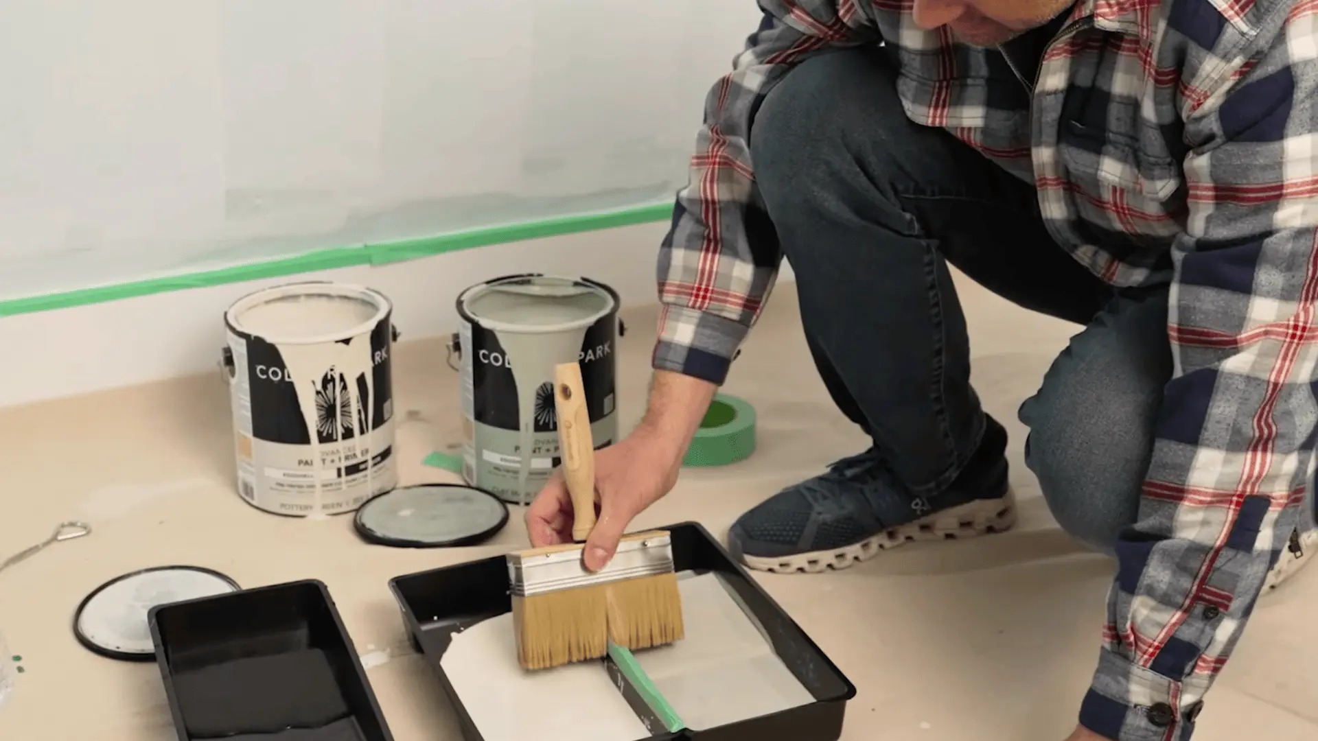 a person loading a stiff bristle brush with limewash paint from a tray beside two colour spark paint cans