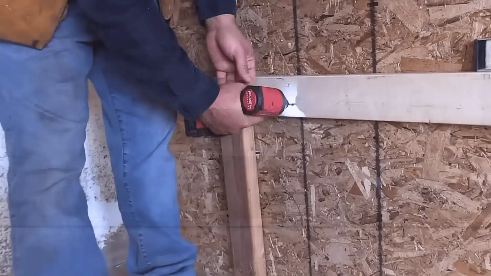 a person mounting a 2x4 wooden support board onto an osb garage wall for diy shelving