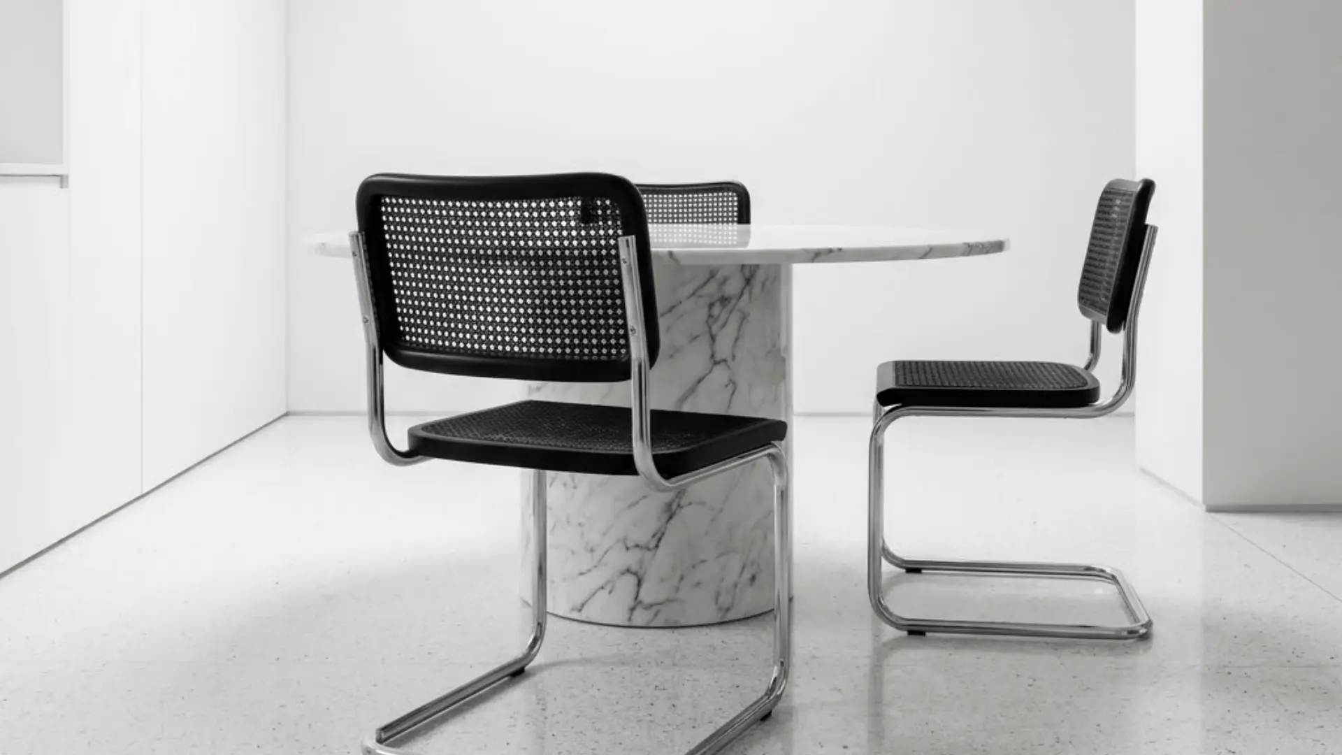 chrome cantilever chair with black cane seat at a marble dining table under a skylight in a minimal white dining room
