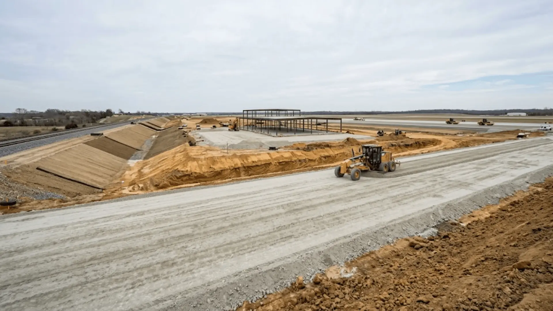 highway subgrade compaction site with parallel railway embankment and building foundation construction in background