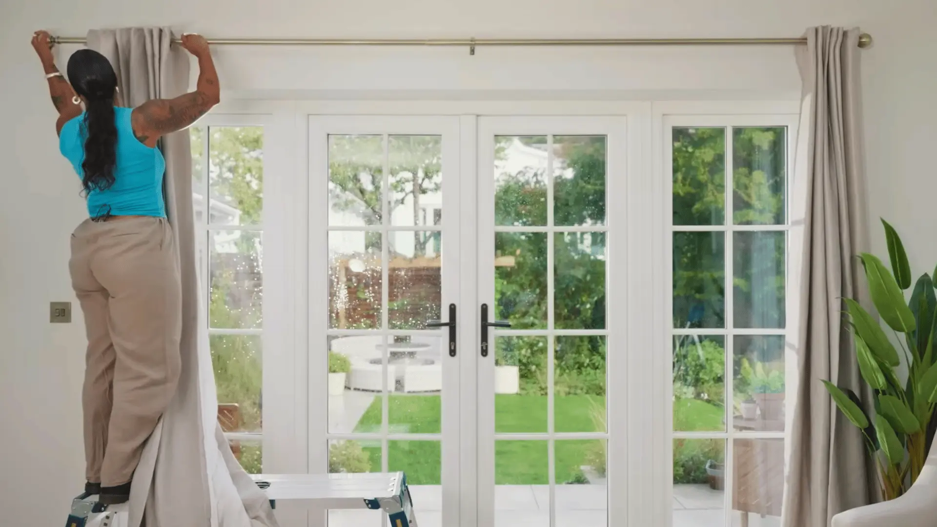 person standing on a ladder threading grommet curtains onto a gold rod above patio doors in a bright living room