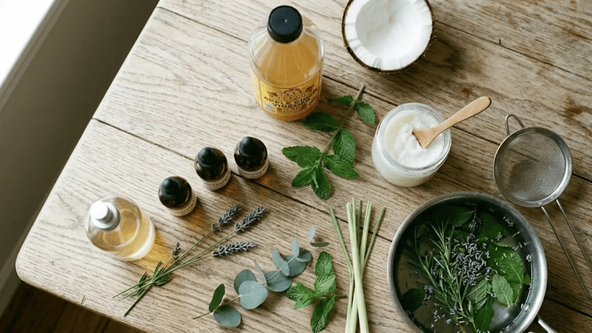 upright essential oil bottles, coconut balm jar, vinegar bottle, and fresh herbs on a wooden table