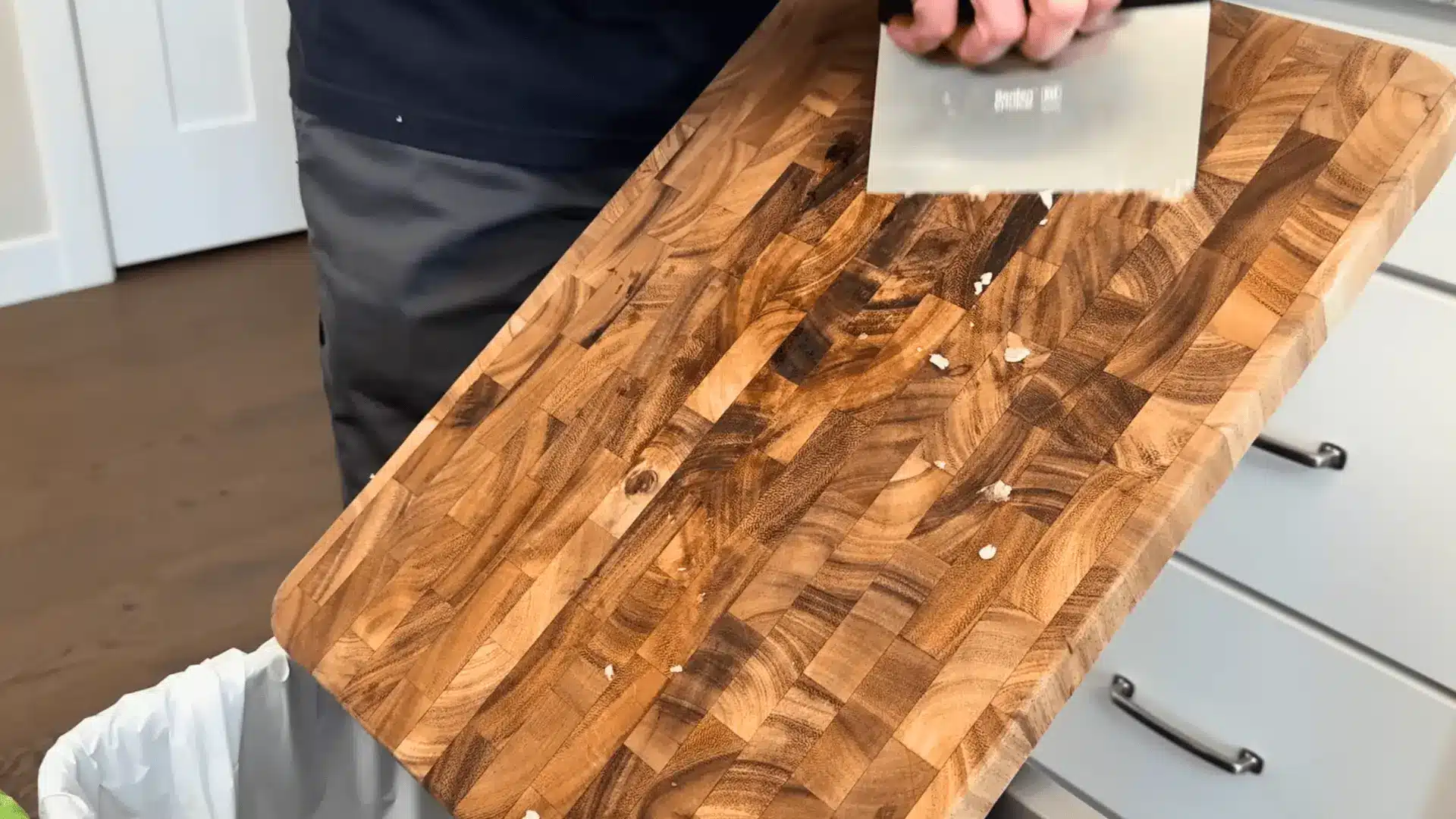using a bench scraper to remove food debris from a wooden cutting board over a trash can