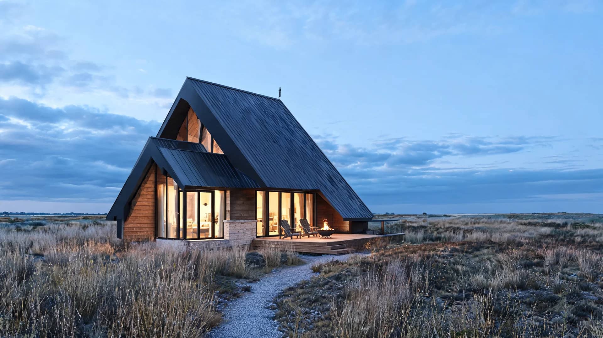 a frame modern farmhouse exterior with dark roof wood cladding and warm glowing glass façade in rural field at dusk