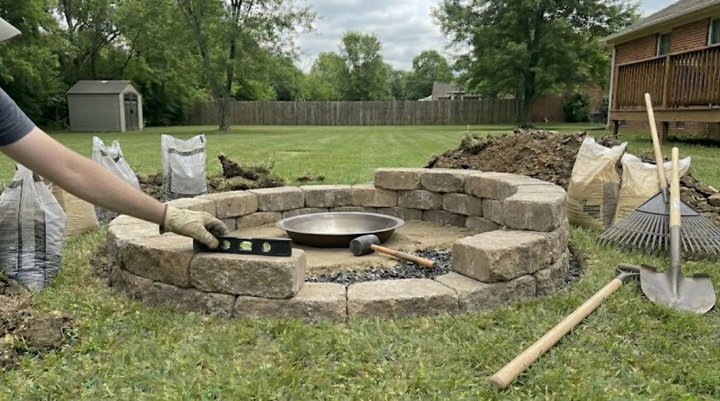 diy fire pit block wall being leveled in backyard with stone pavers, metal ring, mallet, shovel, rake, and supplies