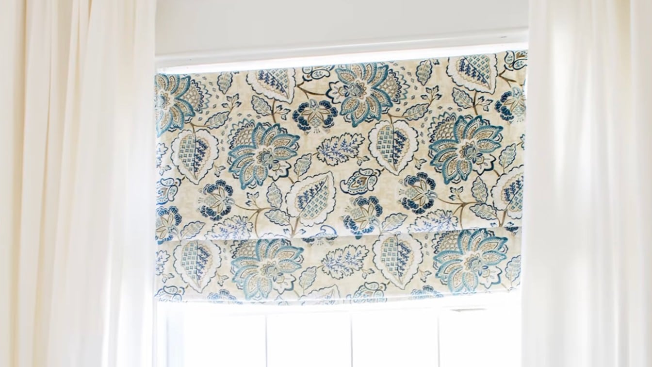 finished patterned roman shade hanging inside a bright window with white curtains and soft light