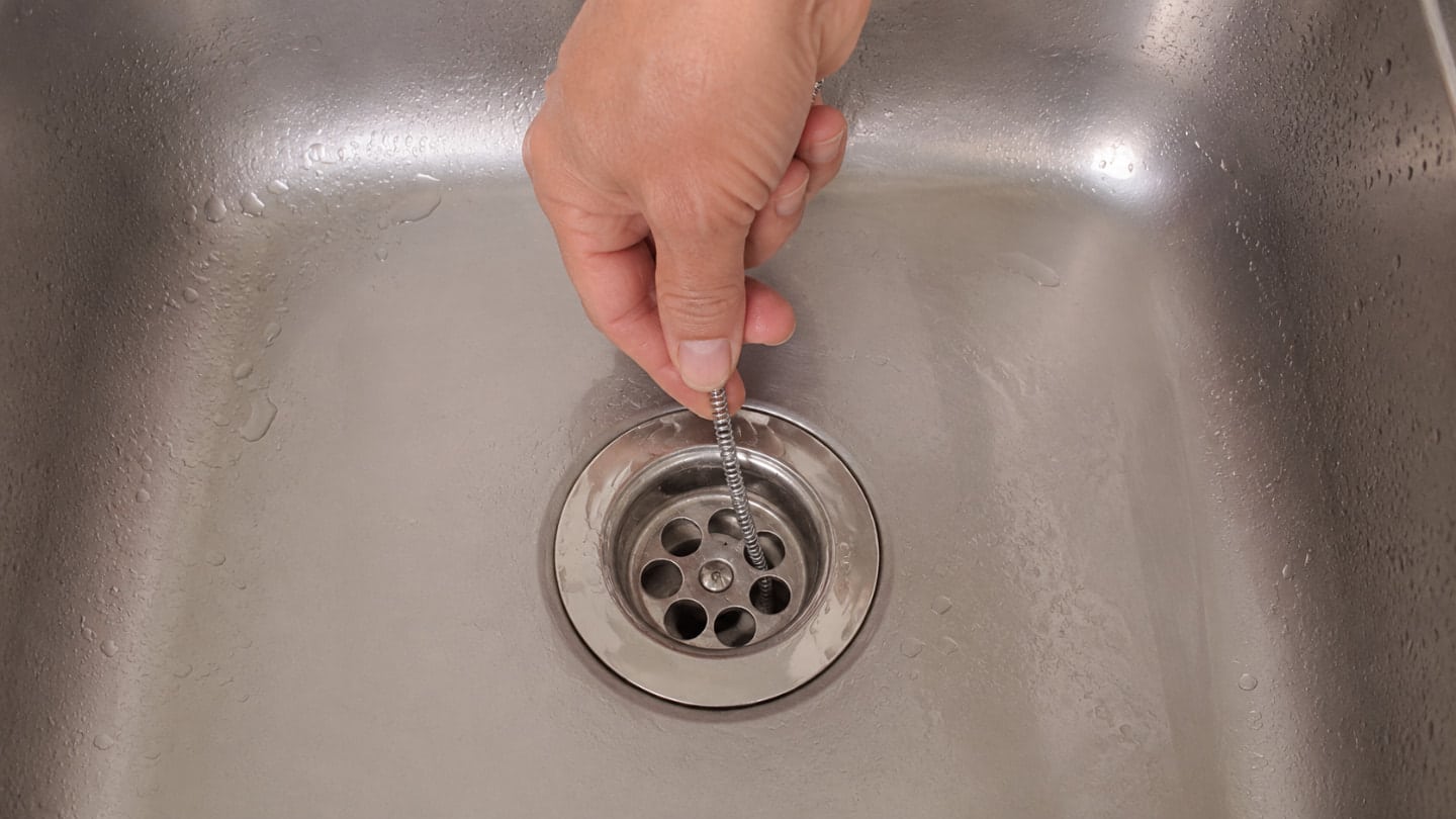 kitchen sink clogged as a hand feeds a drain snake directly into the sink drain from above