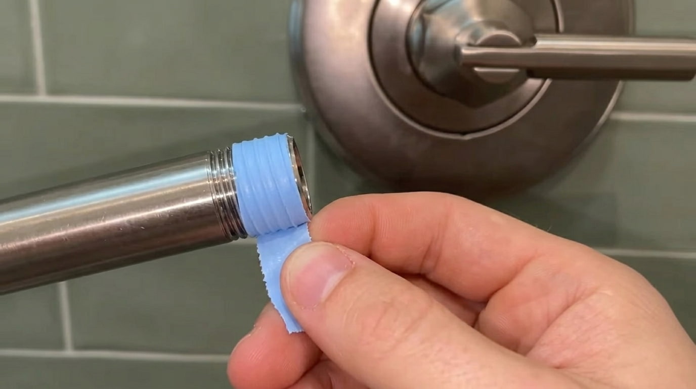 hands wrapping blue plumber tape around metal shower pipe threads against green wall tile
