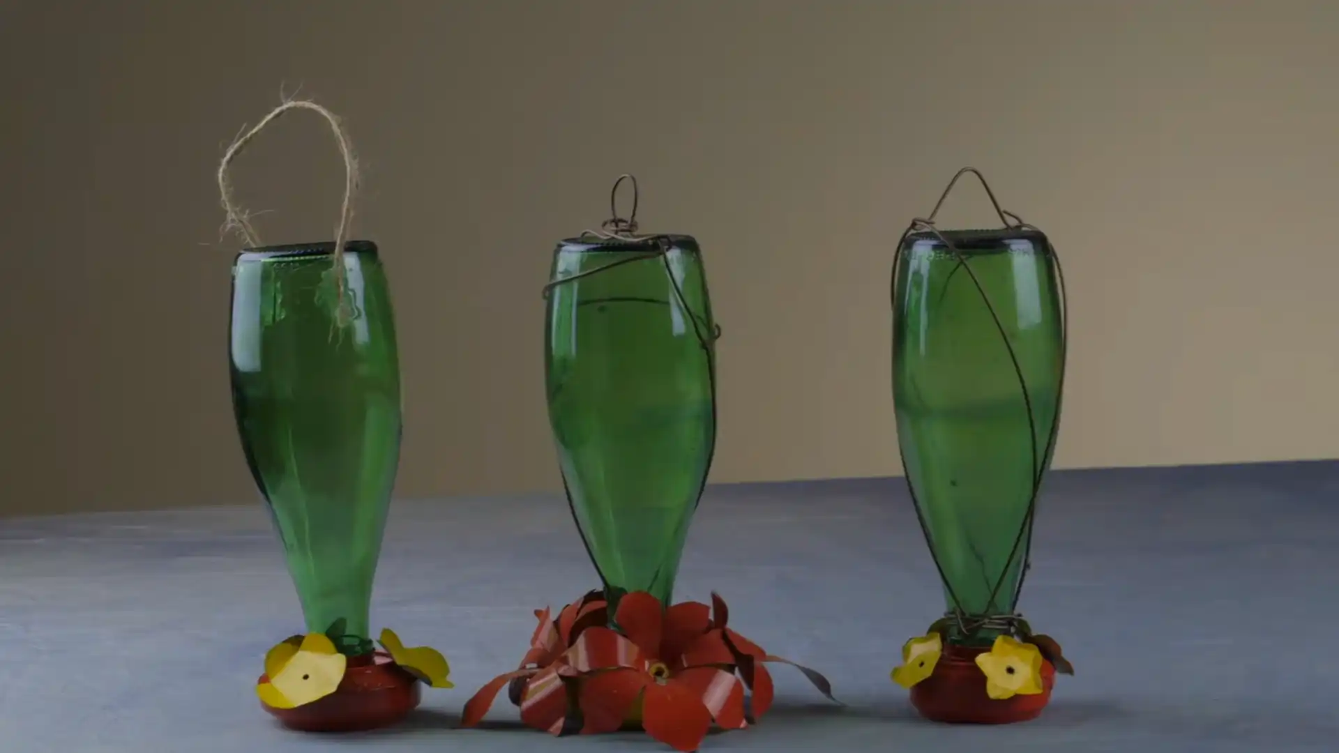 three handmade hummingbird feeders made from green bottles with colorful flower bases displayed upright