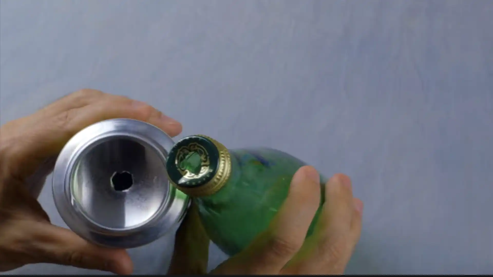 hand holding a soda can with a hole in the bottom beside a capped green glass bottle on a gray surface