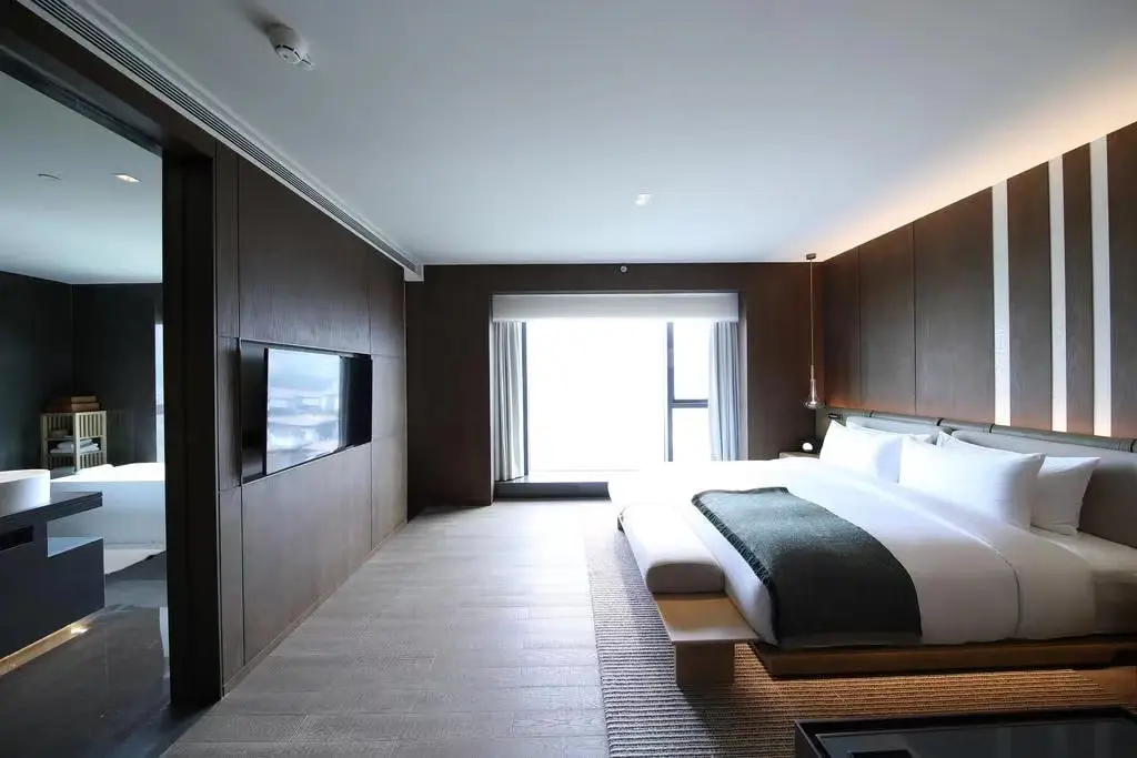 Modern hotel room with large bed, wall-mounted TV, and adjacent bathroom in warm lighting