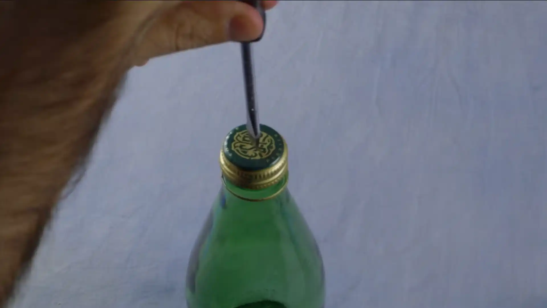 hand using a screwdriver to open the cap of a green glass bottle on a light background
