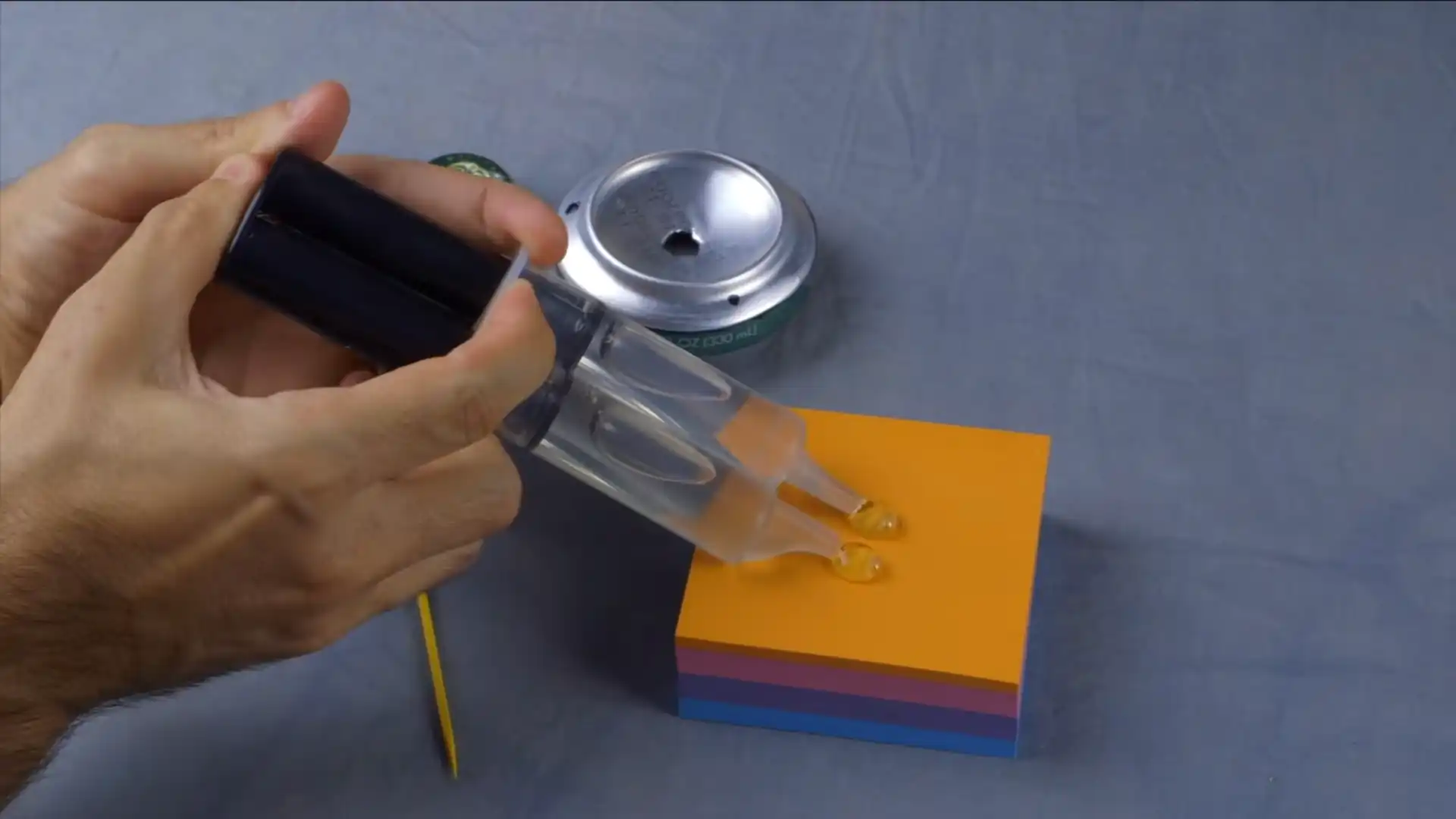 hand applying adhesive from a dual syringe near a cut beverage can and colorful sticky note pad