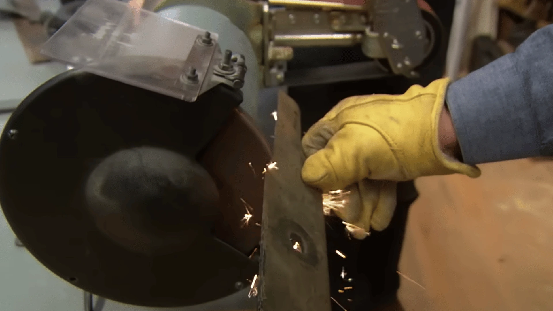 gloved hand sharpens a lawn mower blade on a bench grinder as small sparks fly in a workshop