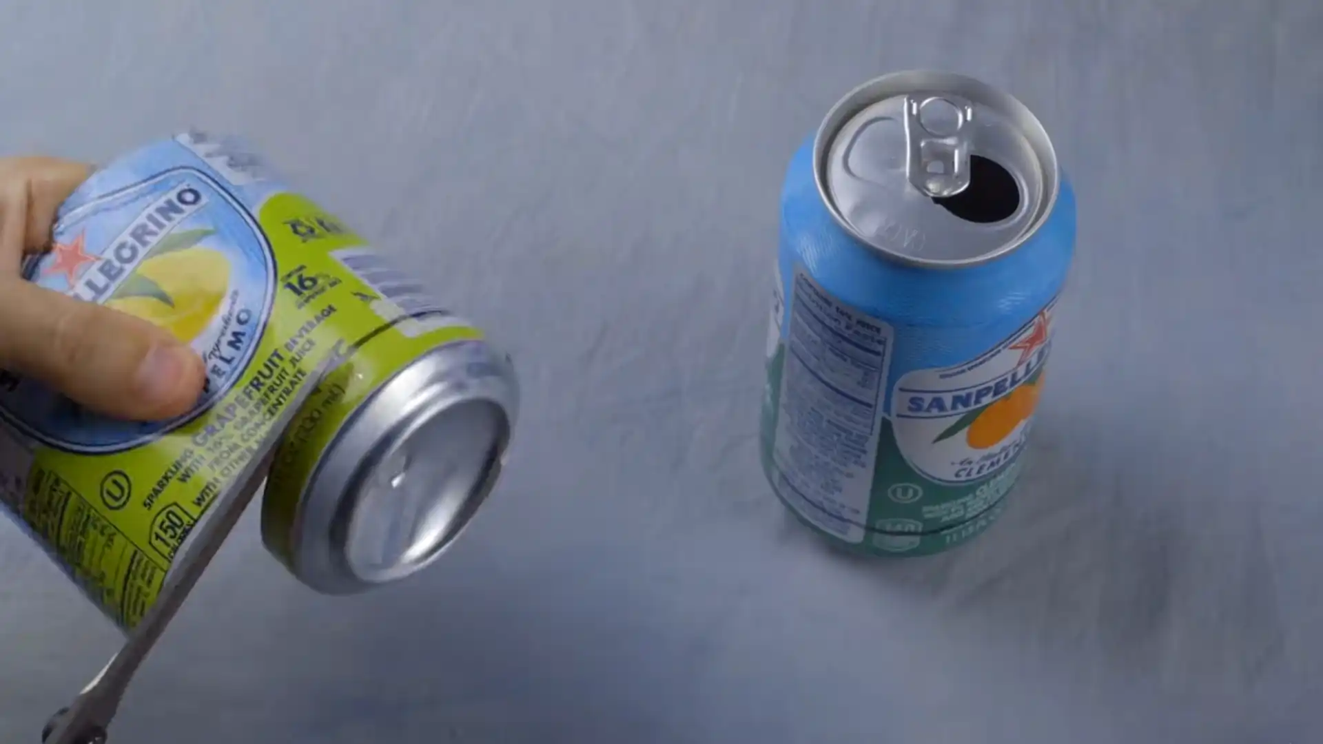 hand holding a soda can beside an opened beverage can on a light gray surface with a metal tool nearby