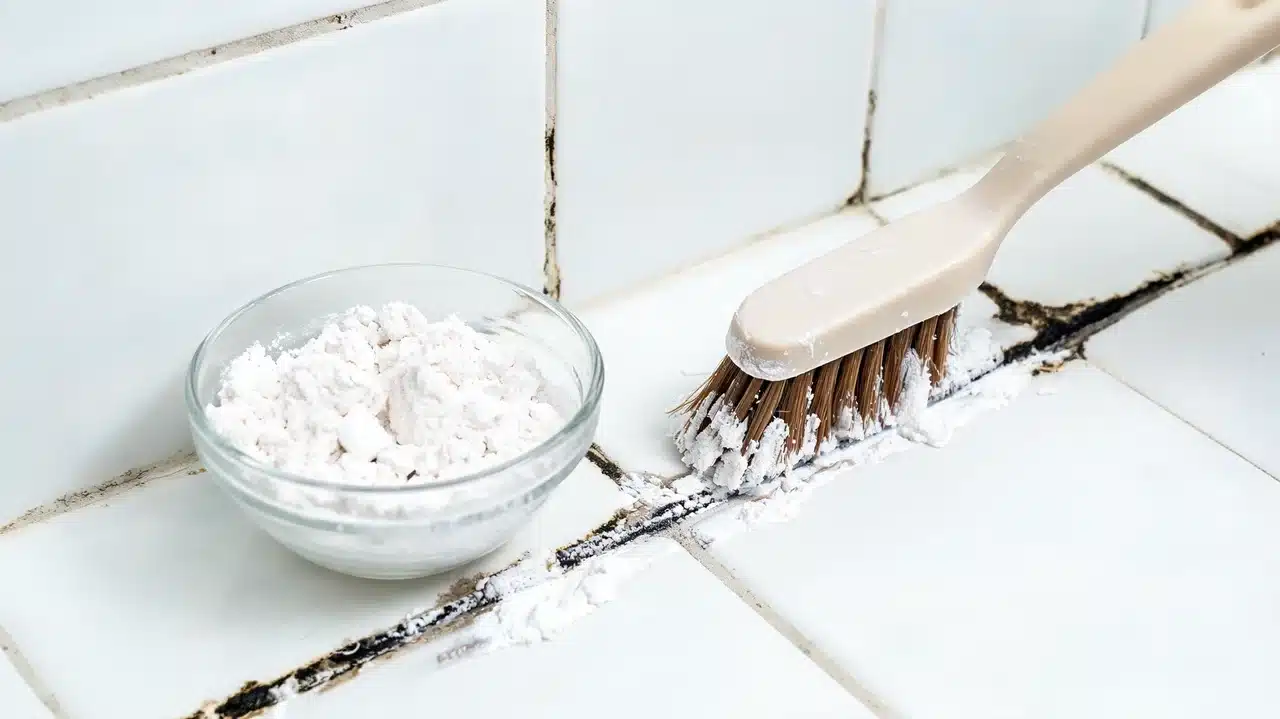 a scrub brush applying white baking soda paste onto moldy black grout lines between white bathroom tiles