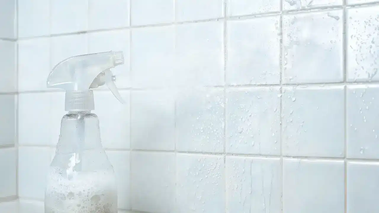 a clear spray bottle beside a foamy cleaning solution freshly misted onto wet white bathroom shower tiles