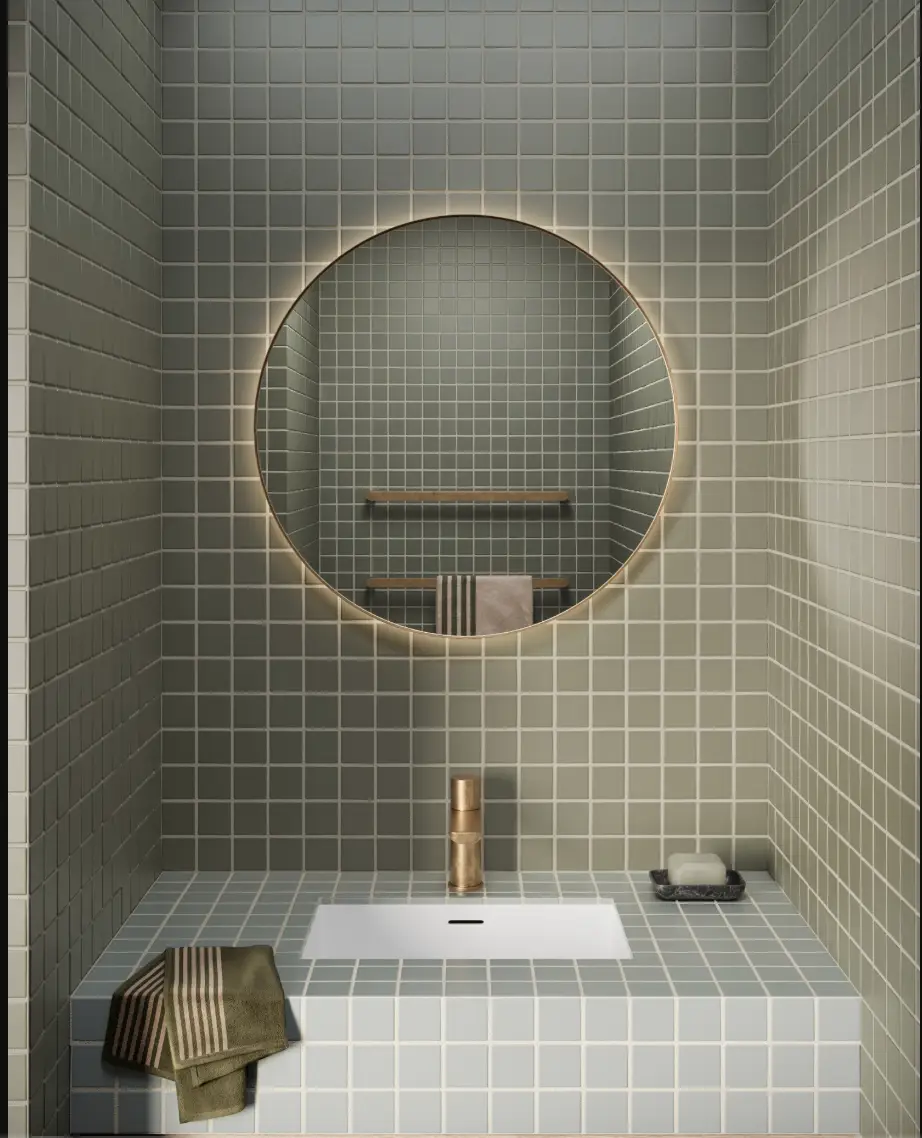 Circular mirror above tiled bathroom sink with brass faucet and folded towels